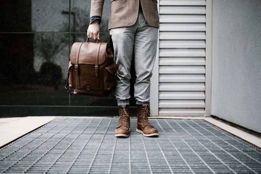 Stylish man wearing brown leather boots, gray pants, and a brown blazer holding a leather backpack — modern men’s clothing and apparel fashion.