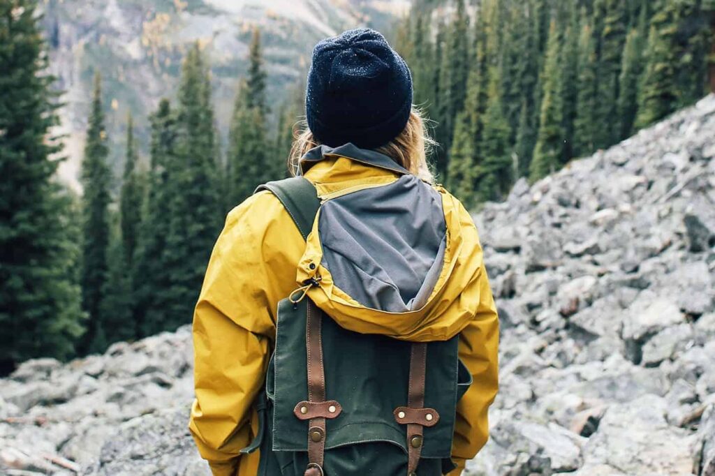 Person wearing a yellow jacket and green backpack hiking through a rocky mountain trail surrounded by pine trees — hiking and backpacking adventure.