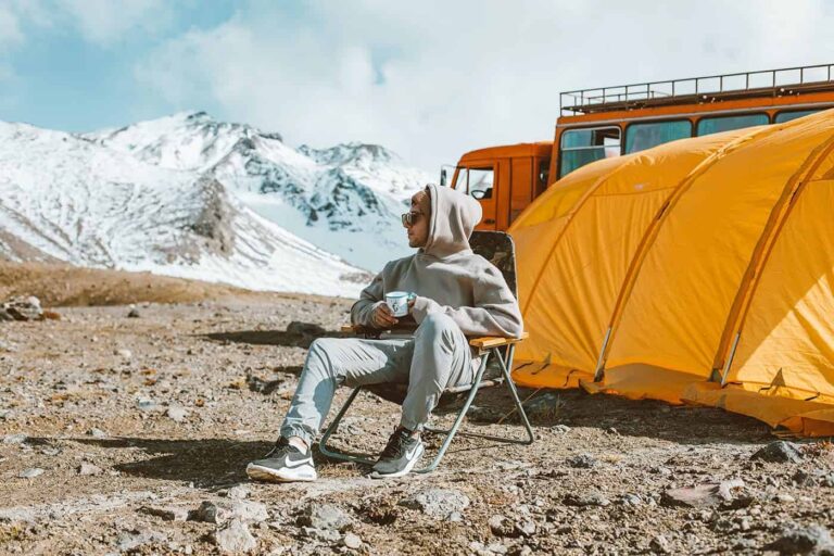 Person relaxing in a camping chair with a mug, sitting beside a bright yellow tent and an orange expedition vehicle, with snow-covered mountains in the background